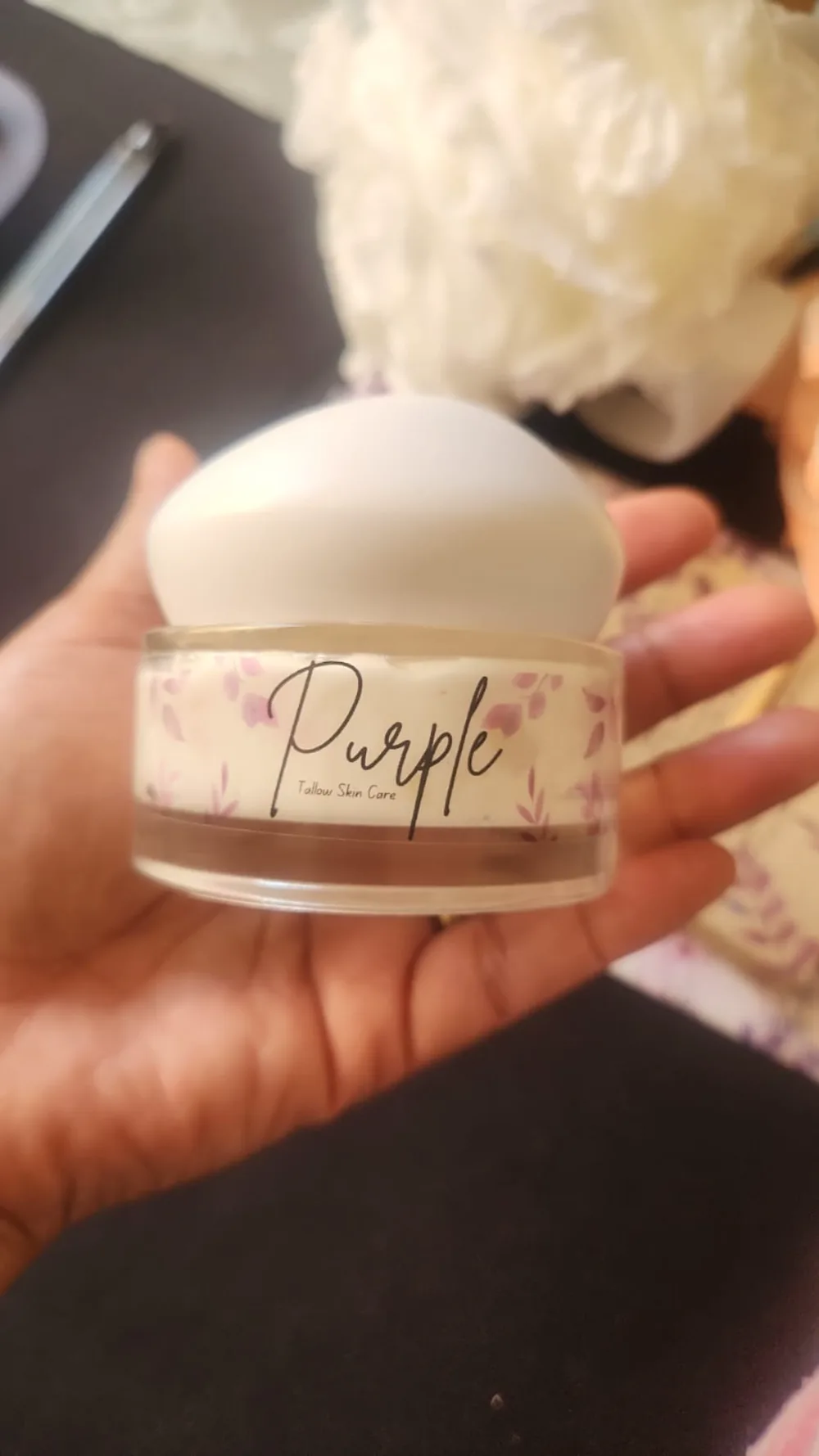 A wide, shallow clear jar of Purple Tallow Skin Care held in one hand, with a domed white lid and a floral label wrapping the whole way round.